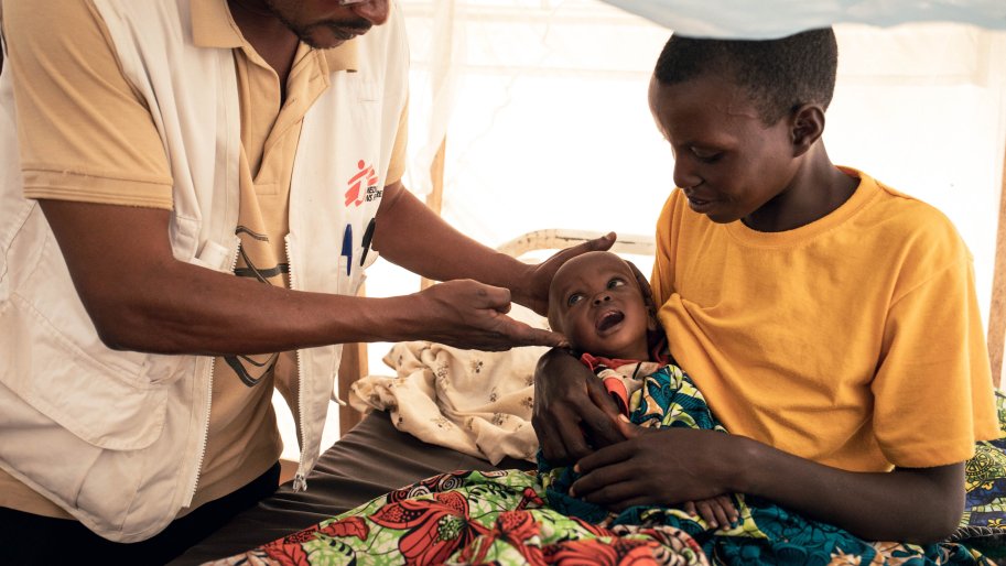 Un médecin de MSF examine Imani, âgée de 9 mois, qui a été admise à l'unité de nutrition thérapeutique intensive pour malnutrition aiguë sévère au poste de santé de MSF situé dans le camp de déplacés de Plaine Savo. Imani est assise sur les genoux de sa mère, Nehema, âgée de 20 ans, qui la tient dans ses bras pendant l'examen. 