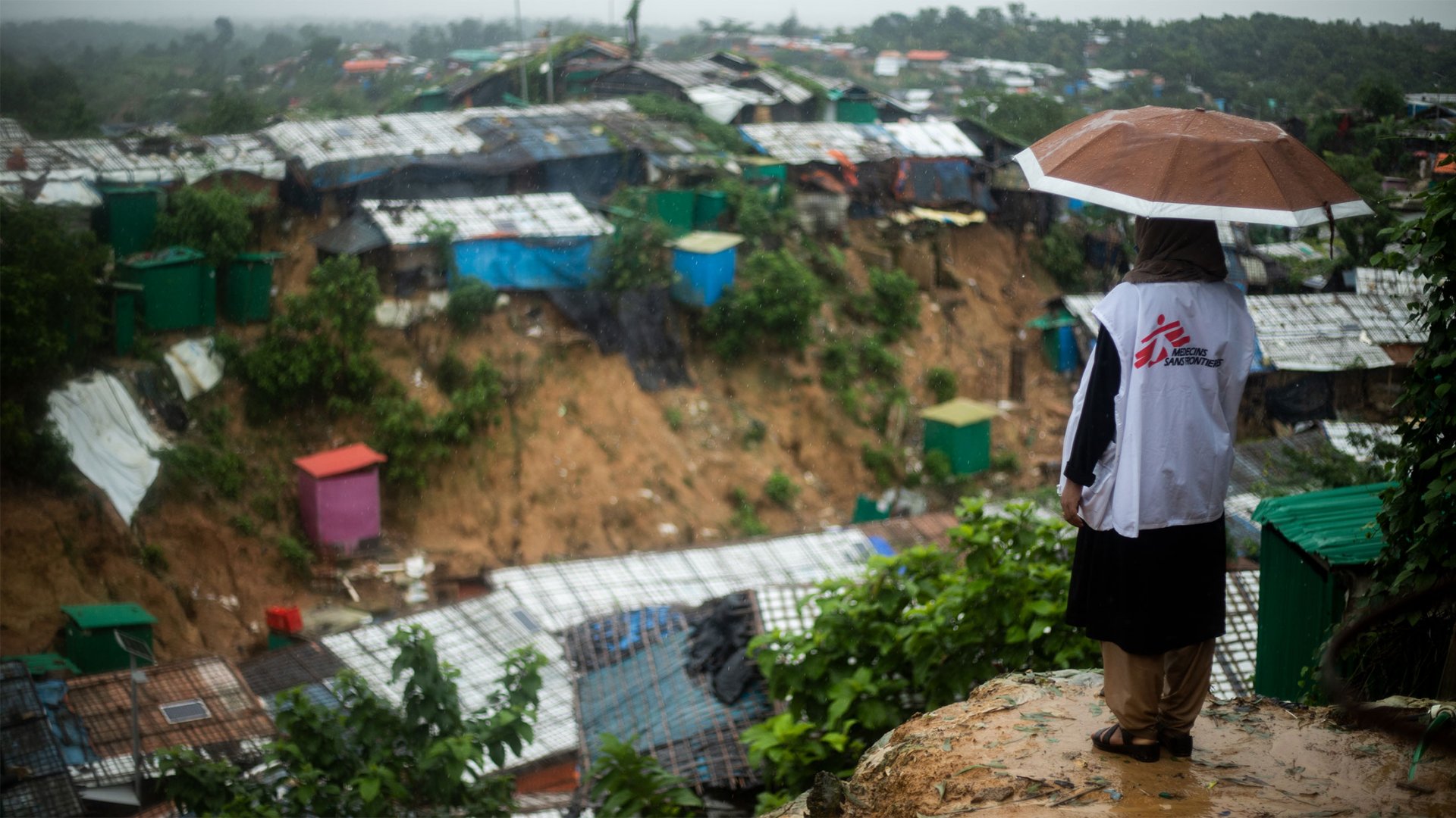 Eine Person von MSF schaut bei Regen von unten auf das Cox’s Bazar Kamp, dabei trägt sie einen Regenschirm.