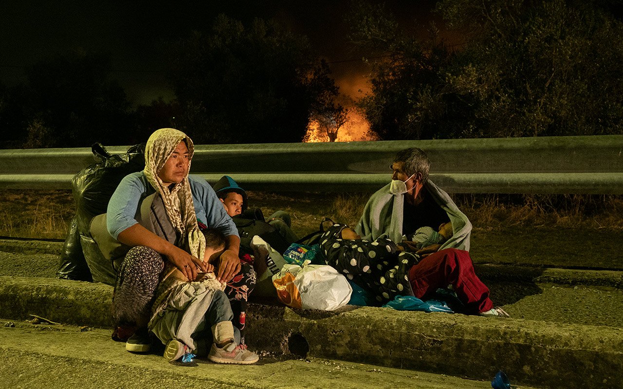 Deux parents et trois enfants, assis sur un trottoir, regardent au loin les flammes dévorer Moria