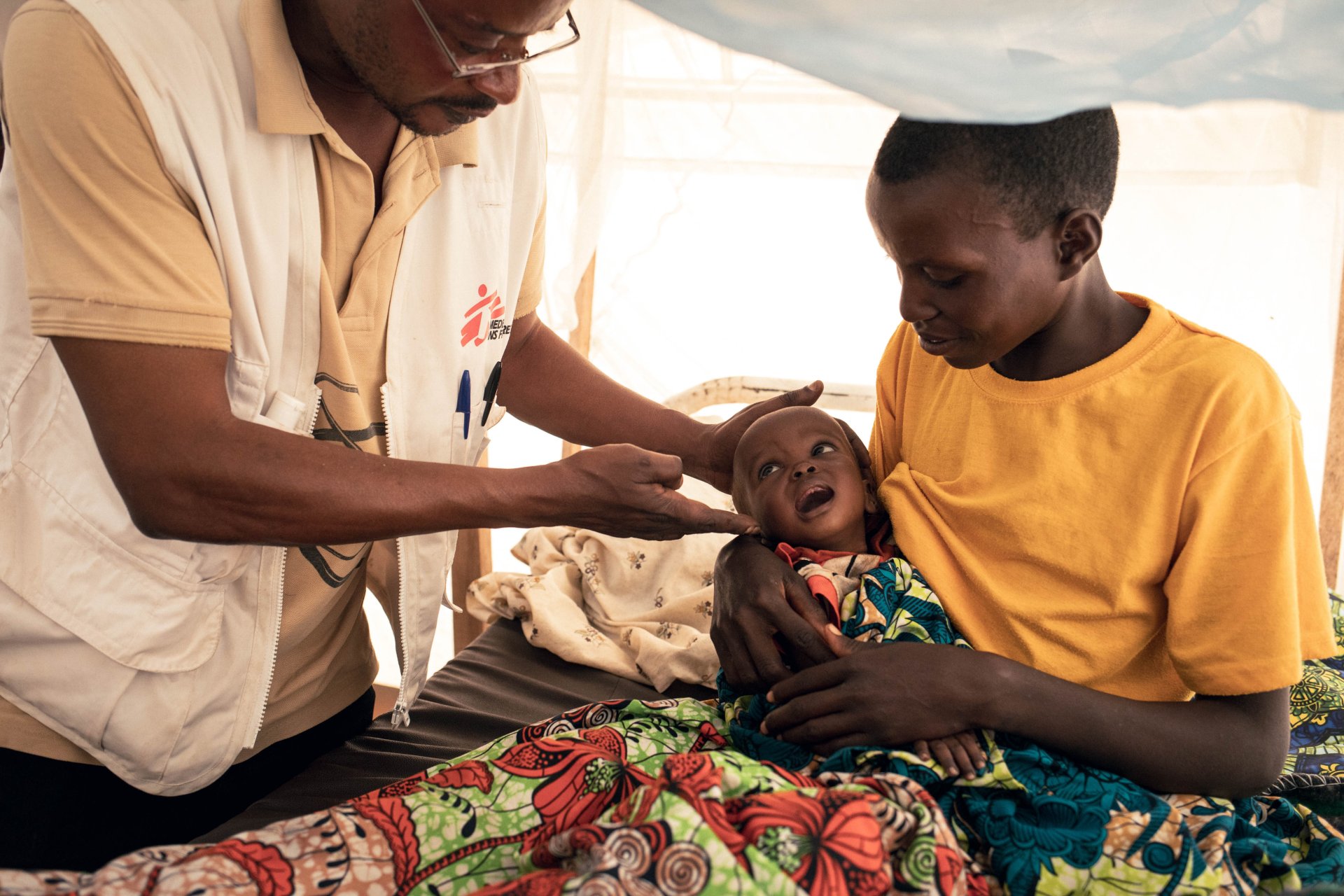 Un médecin de MSF examine Imani, âgée de 9 mois, qui a été admise à l'unité de nutrition thérapeutique intensive pour malnutrition aiguë sévère au poste de santé de MSF situé dans le camp de déplacés de Plaine Savo. Imani est assise sur les genoux de sa mère, Nehema, âgée de 20 ans, qui la tient dans ses bras pendant l'examen. 