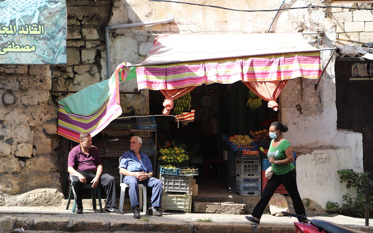 2 hommes assis sur des chaises dans une rues de Beyrouth discutent. Une femme avec un masque chirurgical marche sur le trottoir
