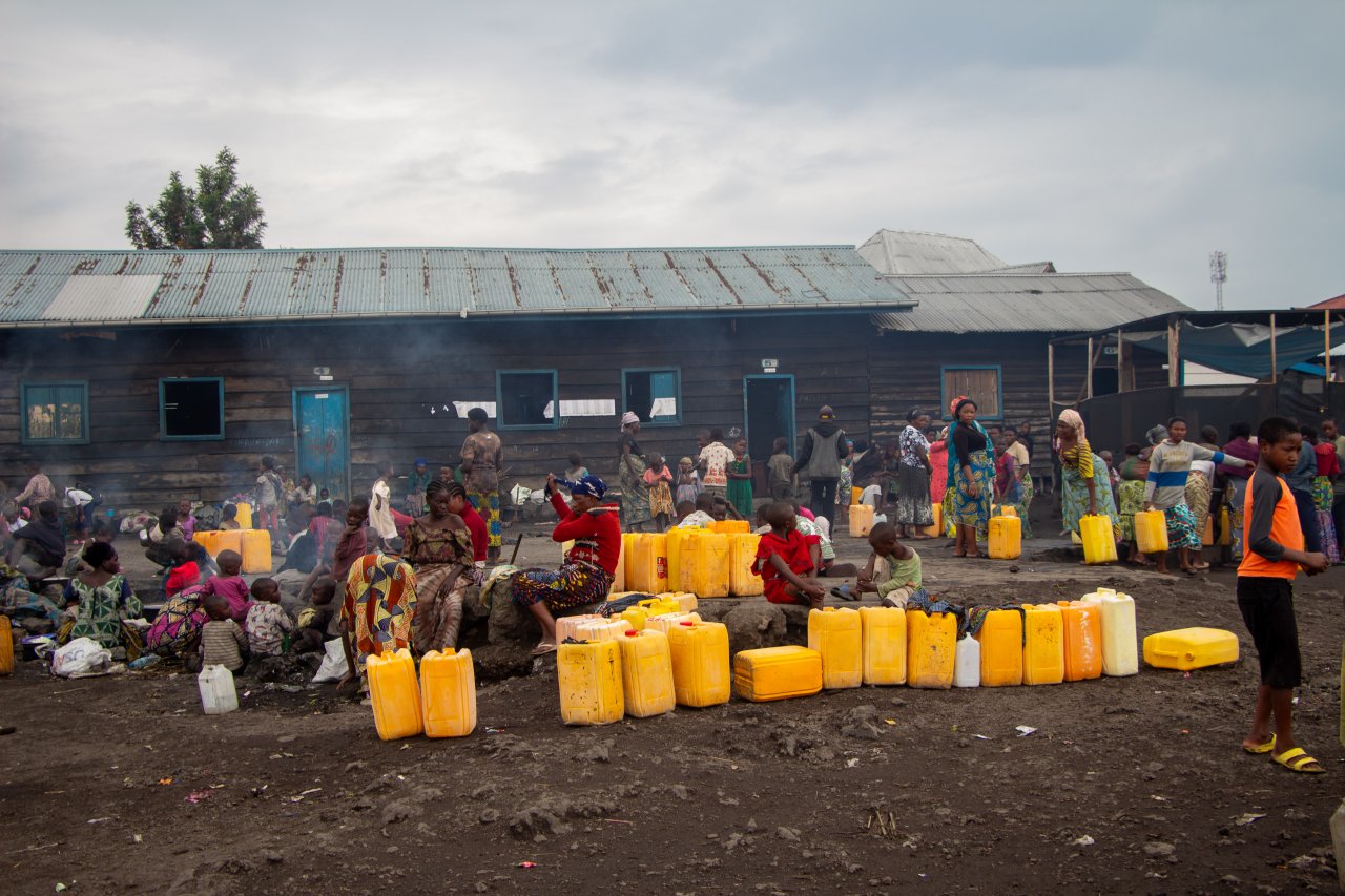 Die vertriebenen Menschen verbringen viel Zeit mit Warten, bis der Lastwagen mit Trinkwasser ankommt. Meistens sind es die Grossmütter und Kinder, die auf die Lieferung des Wassers warten. Sobald der Lastwagen vor Ort ist, ist es die Aufgabe der jüngeren Frauen, die Kanister zu füllen und sie zu ihren Unterkünften zu bringen. DR Kongo, Febreuar 2024.