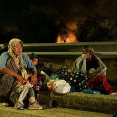 Deux parents et trois enfants, assis sur un trottoir, regardent au loin les flammes dévorer Moria