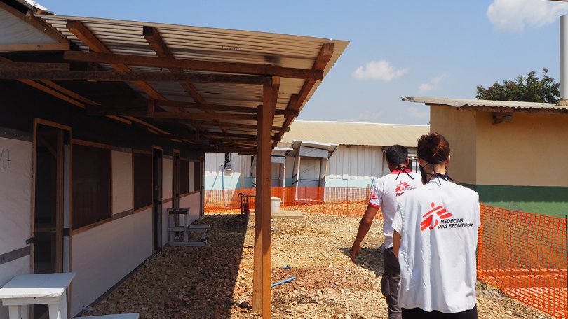2 staffs en gilets MSF marchent entre une barrière orange et une petite maison en bois et en tôle. 